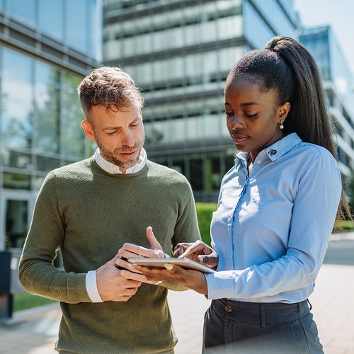 Two professionals, a man and a woman, are engaged in a discussion while examining a tablet. They are outdoors near modern office buildings, under clear skies.