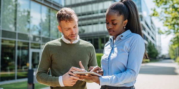 Two professionals, a man and a woman, are engaged in a discussion while examining a tablet. They are outdoors near modern office buildings, under clear skies.