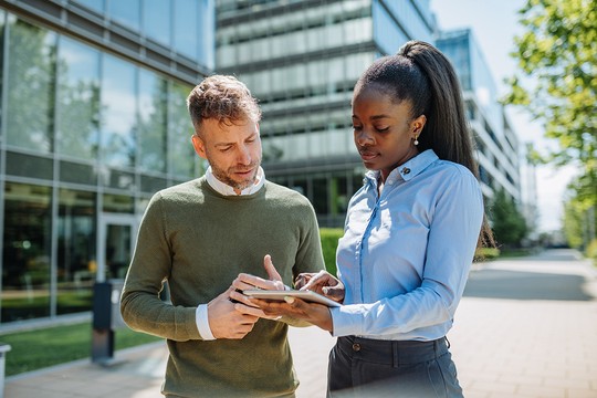 Two professionals, a man and a woman, are engaged in a discussion while examining a tablet. They are outdoors near modern office buildings, under clear skies.