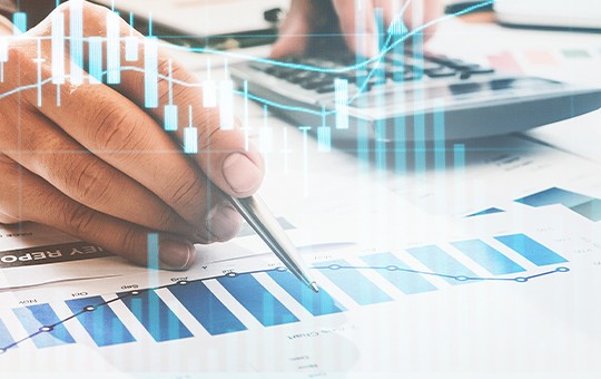 A hand holds a pen while analyzing colorful bar graphs and charts on paper. A calculator rests nearby on a desk, suggesting a focused data analysis context.