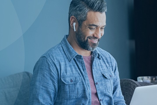 A man wearing earbuds smiles while using a laptop. He sits on a couch in a room with dark walls and a bookshelf in the background filled with books and decorative items.