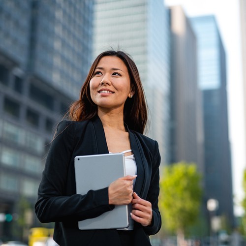 A woman in a black blazer holds a tablet and smiles confidently. She stands amidst tall modern buildings and greenery, suggesting a vibrant urban environment.