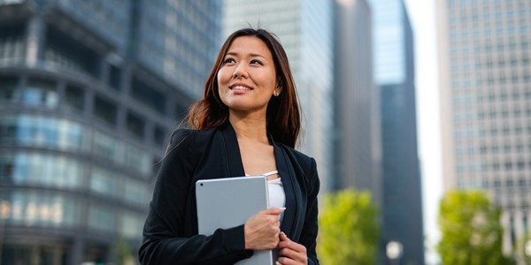 A woman in a black blazer holds a tablet and smiles confidently. She stands amidst tall modern buildings and greenery, suggesting a vibrant urban environment.