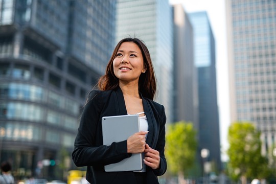A woman in a black blazer holds a tablet and smiles confidently. She stands amidst tall modern buildings and greenery, suggesting a vibrant urban environment.