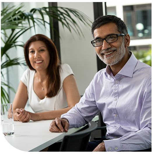 Two professionals sit at a modern table in a bright room with large windows. One man, wearing glasses and a light purple shirt, smiles, while a woman in a white top appears engaged. A glass of water is on the table.