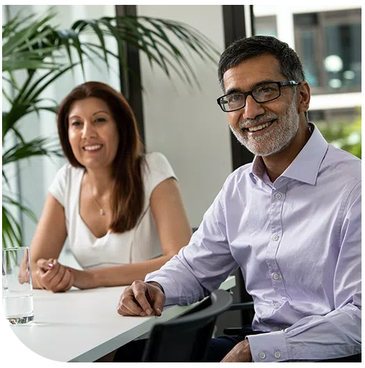 Two professionals sit at a modern table in a bright room with large windows. One man, wearing glasses and a light purple shirt, smiles, while a woman in a white top appears engaged. A glass of water is on the table.