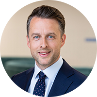 A man with short, styled hair smiles confidently while wearing a formal dark suit and a polka-dot tie, standing in an office environment featuring soft seating in the background.