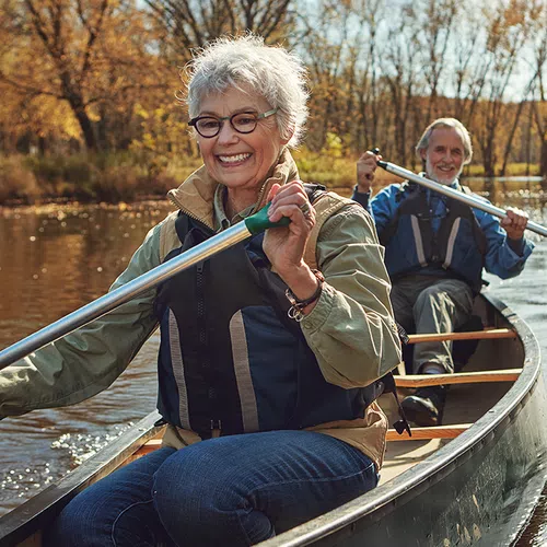 A couple joyfully paddles a canoe on a calm river surrounded by autumn trees. The woman in front smiles broadly, while the man in the back maintains a cheerful expression.