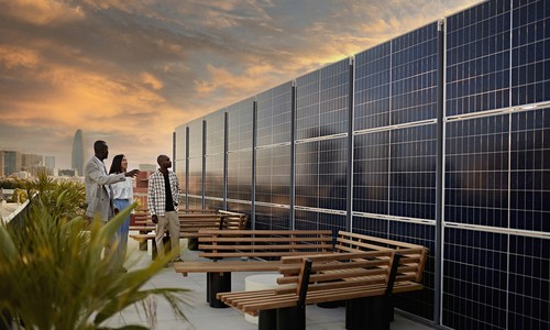 A group of three people observes solar panels on a rooftop, discussing among themselves. The scene is set against a dramatic sunset, with city buildings in the background and benches nearby.