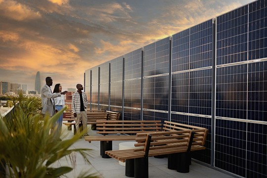 A group of three people observes solar panels on a rooftop, discussing among themselves. The scene is set against a dramatic sunset, with city buildings in the background and benches nearby.