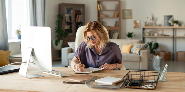 A woman writes in a notebook at a wooden desk in a well-lit home office, surrounded by bookshelves, plants, and a comfortable couch, suggesting a productive and inviting workspace.