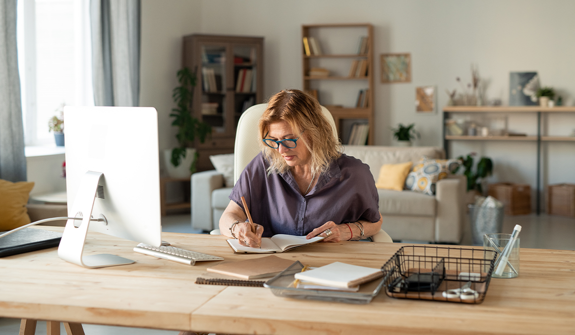 A woman writes in a notebook at a wooden desk in a well-lit home office, surrounded by bookshelves, plants, and a comfortable couch, suggesting a productive and inviting workspace.