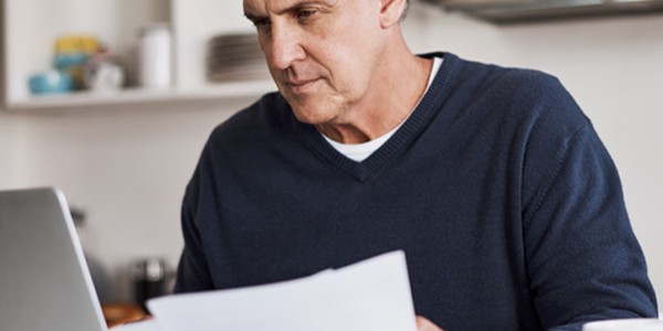 A middle-aged man sits at a kitchen counter, focused on a laptop while holding printed documents, surrounded by kitchen utensils and a white mug, conveying a productive environment.