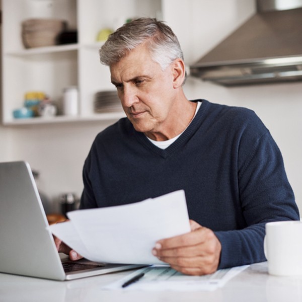 A middle-aged man sits at a kitchen counter, focused on a laptop while holding printed documents, surrounded by kitchen utensils and a white mug, conveying a productive environment.