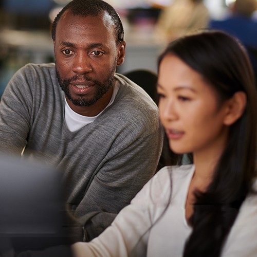 A man and a woman are seated at a desk, collaboratively reviewing a computer screen. The background shows an office environment with people working and blurred elements indicating a busy space.