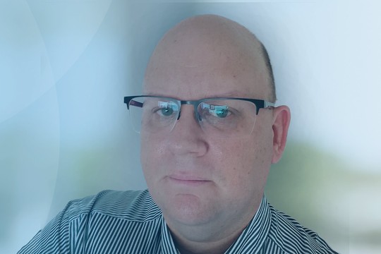 A man with glasses gazes directly while partially smiling. He is dressed in a striped shirt, with a soft blue background providing a calm atmosphere.