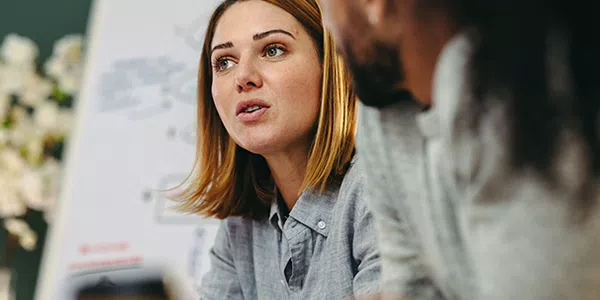 A woman with shoulder-length hair speaks intently during a meeting, while a man listens nearby. A whiteboard with diagrams is positioned in the background, indicating a collaborative workspace.