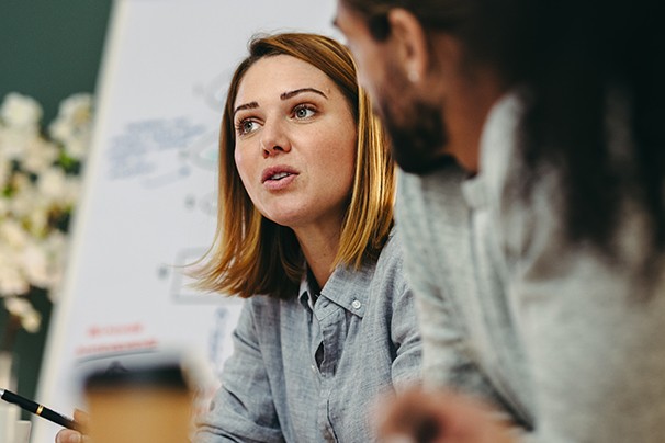 A woman with shoulder-length hair speaks intently during a meeting, while a man listens nearby. A whiteboard with diagrams is positioned in the background, indicating a collaborative workspace.