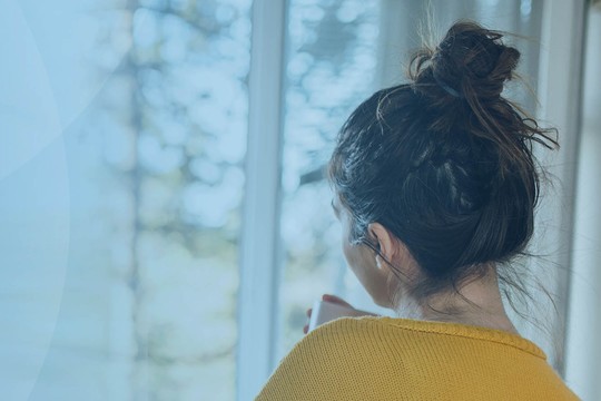 A woman with her back to the viewer gazes out a window, holding a phone, while surrounded by a softly lit interior and trees visible in the background.