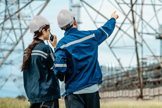 Two workers in safety helmets communicate with a radio while observing power lines. One gestures toward the structures, set against a cloudy sky and grassy surroundings.