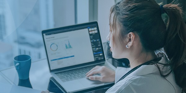 A woman in a white coat is working on a laptop, reviewing graphs and data. A blue cup sits beside her, and she appears focused in a modern office environment.