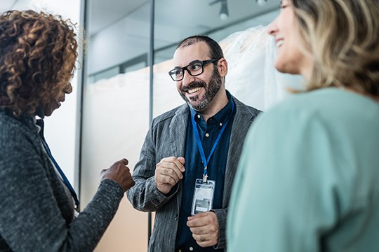 Three professionals engage in a friendly conversation, smiling and gesturing. They stand in a modern, well-lit setting with glass walls, suggesting a collaborative or workplace environment.