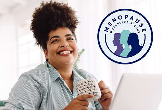 A woman with curly hair smiles while holding a mug, sitting at a desk. Natural light filters through the window, and a logo featuring silhouettes of women and text reads “MENOPAUSE WORKPLACE PLEDGE.”