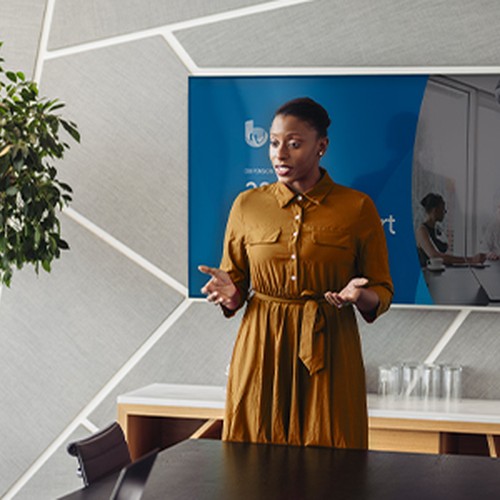 A woman in a brown dress stands confidently in front of a group, presenting to them while a television displays a meeting scene. The setting features a modern conference room with plants.