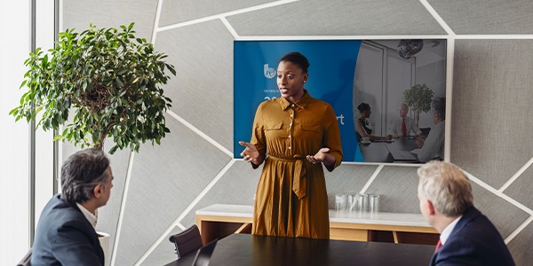 A woman in a brown dress stands confidently in front of a group, presenting to them while a television displays a meeting scene. The setting features a modern conference room with plants.