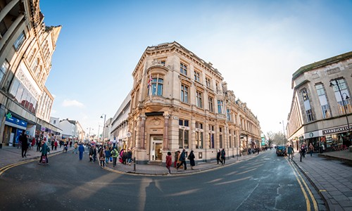 A historic building with ornate architecture stands on a bustling street corner. Pedestrians stroll by while shops line the street, under a clear blue sky.