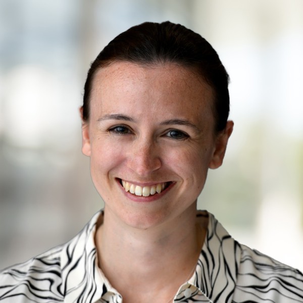 A smiling woman with dark hair pulled back wears a black-and-white striped shirt. She stands in a blurred, bright environment, projecting a friendly and approachable demeanor.