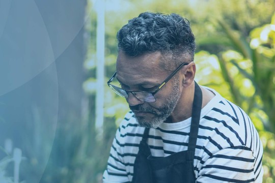 A man with curly hair, wearing glasses and a striped shirt, tends to plants in a garden filled with greenery and colorful flowers, focusing intently on his work.