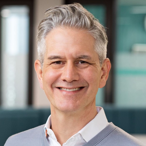 A middle-aged man with short, silver hair smiles warmly, wearing a light grey sweater over a white shirt. He's positioned in a modern, well-lit office environment with soft, muted colors in the background.