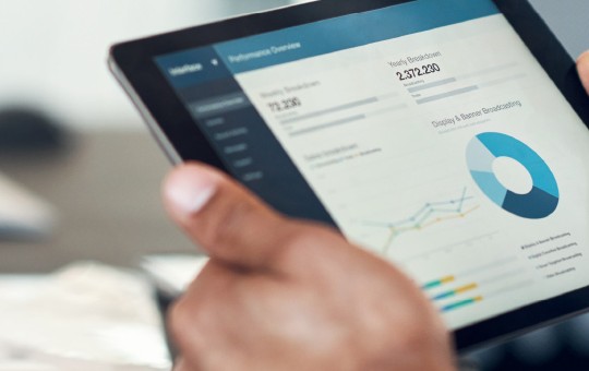 A person is holding a tablet displaying detailed data analytics, including graphs and a pie chart, in a modern office environment with blurred office items in the background.
