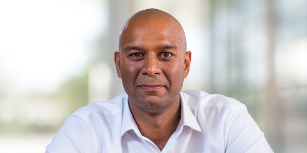 A man with a bald head and a serious expression sits at a table. He wears a white shirt and is positioned in a bright, blurred indoor setting.