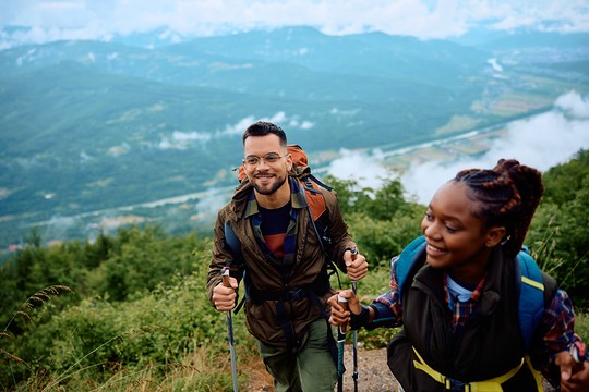 Two hikers are ascending a scenic mountain trail, smiling and holding trekking poles. Lush greenery surrounds them, and a valley is visible below, with clouds and mountains in the distance.