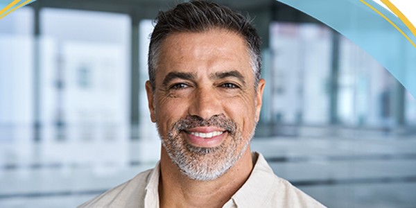 A man with salt-and-pepper hair and a beard smiles warmly. He is wearing a light-colored shirt, positioned in a bright, modern office environment with blurred furniture in the background.