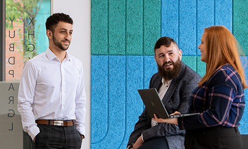 Three professionals engage in discussion. One man stands while two others sit, with one holding a laptop. The setting features colorful, textured wall panels in a modern office environment.