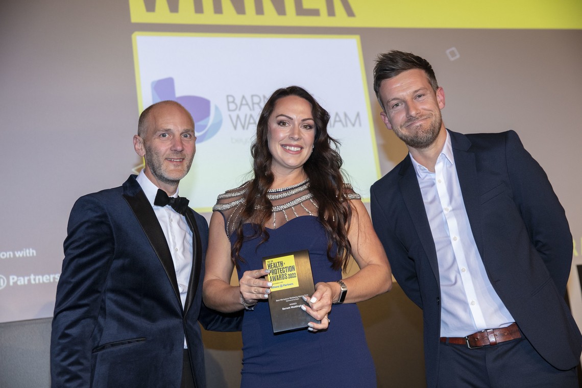 A woman in a formal dark dress holds an award, surrounded by two men in suits. The background displays a presentation screen, indicating a celebratory event.