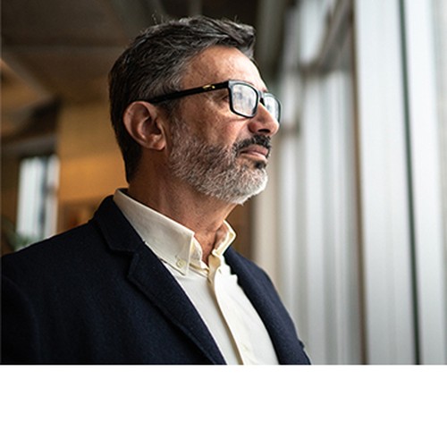 A man with glasses and a beard is gazing thoughtfully out of a large window, standing in a modern indoor environment filled with natural light.