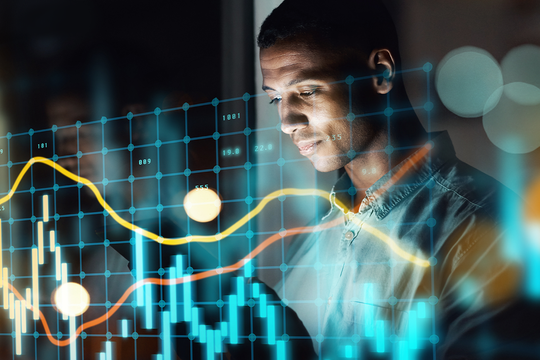 A person examines digital financial data on a transparent screen, highlighting various graphs and lines in bright colors. The environment appears dim, adding focus to the data.