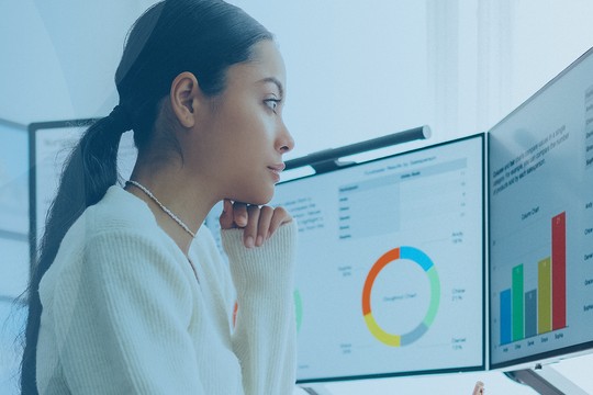A woman in a white sweater focuses intently on dual monitors displaying colorful data visualizations. The setting appears professional, with light filtering through a window in a modern office.