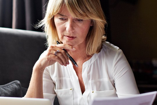 A woman is seated at a table, examining documents closely while occasionally writing notes. She is in a well-lit indoor setting with a sofa and curtains in the background.