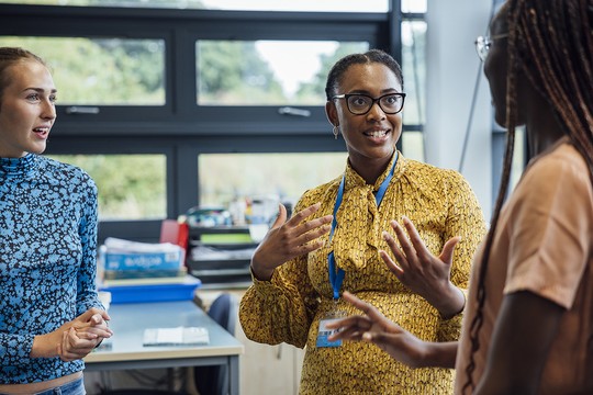 Three women engage in animated conversation indoors, gesturing expressively. A desk with papers and a blue container is nearby, while large windows allow natural light to illuminate the room.