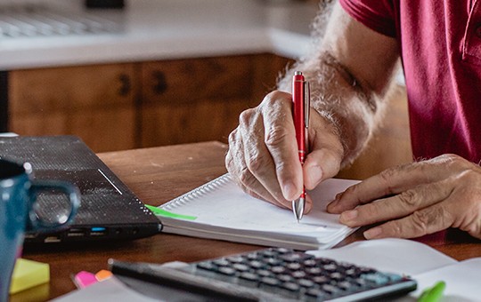 A person is writing in a notebook with a red pen, seated at a desk with a laptop, calculator, coffee mug, and colorful sticky notes, surrounded by a bright, homey kitchen environment.