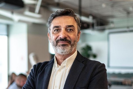 A middle-aged man with a beard stands confidently with arms crossed. He is in a bright, modern office space, featuring large windows and plants, indicating a professional setting.