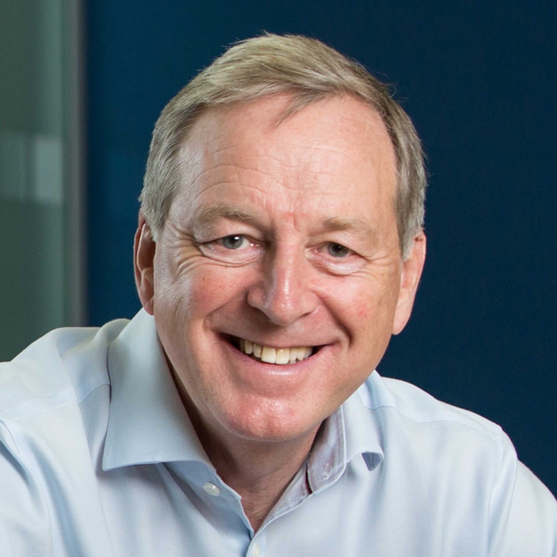 A middle-aged man with short, light gray hair smiles warmly, seated in an indoor office environment with a blue backdrop, conveying a friendly and approachable demeanor.