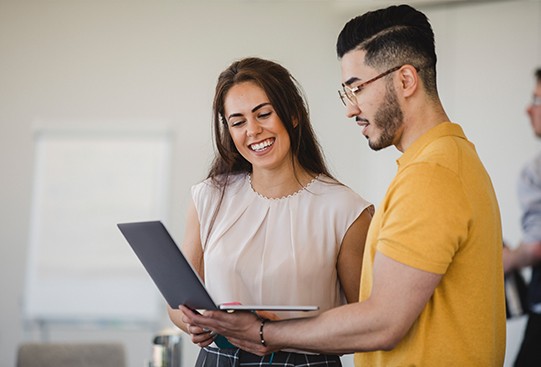 A woman and a man smile while discussing a laptop in a modern office setting. The woman points at the screen, engaged in conversation, with a whiteboard and other people visible in the background.