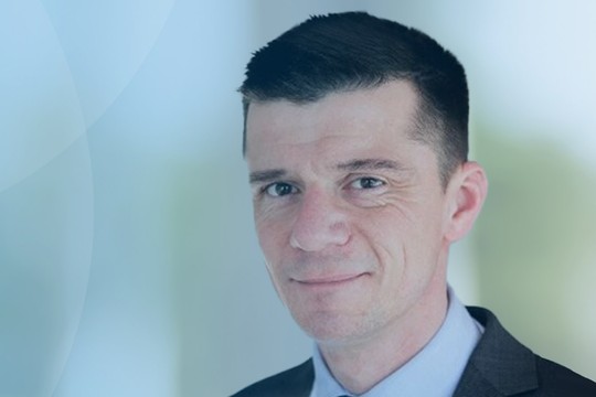 A man in a suit is smiling confidently. He is positioned against a soft blue background with subtle gradients, suggesting a professional or corporate environment.
