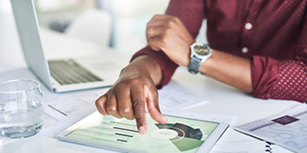 A hand is tapping a touchscreen tablet displaying graphs and data. A laptop, glass of water, and paper documents are on a white table in a bright office environment.